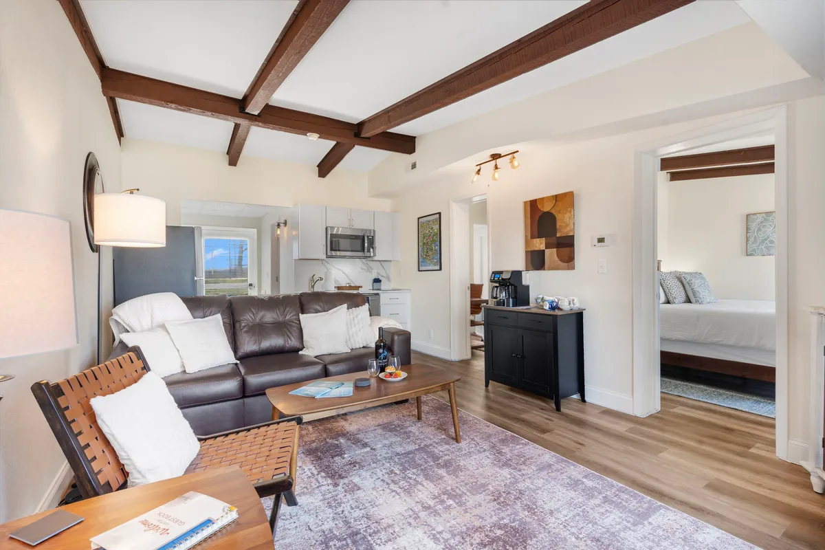 The image showcases a warm, inviting living area with a plush leather sofa, rustic wooden beams overhead, a stylish coffee table, and a modern open kitchen. Artwork on the wall adds character, while the space is bathed in natural light, creating a cozy ambiance perfect for relaxing, socializing, or enjoying a meal after a day by the lake.