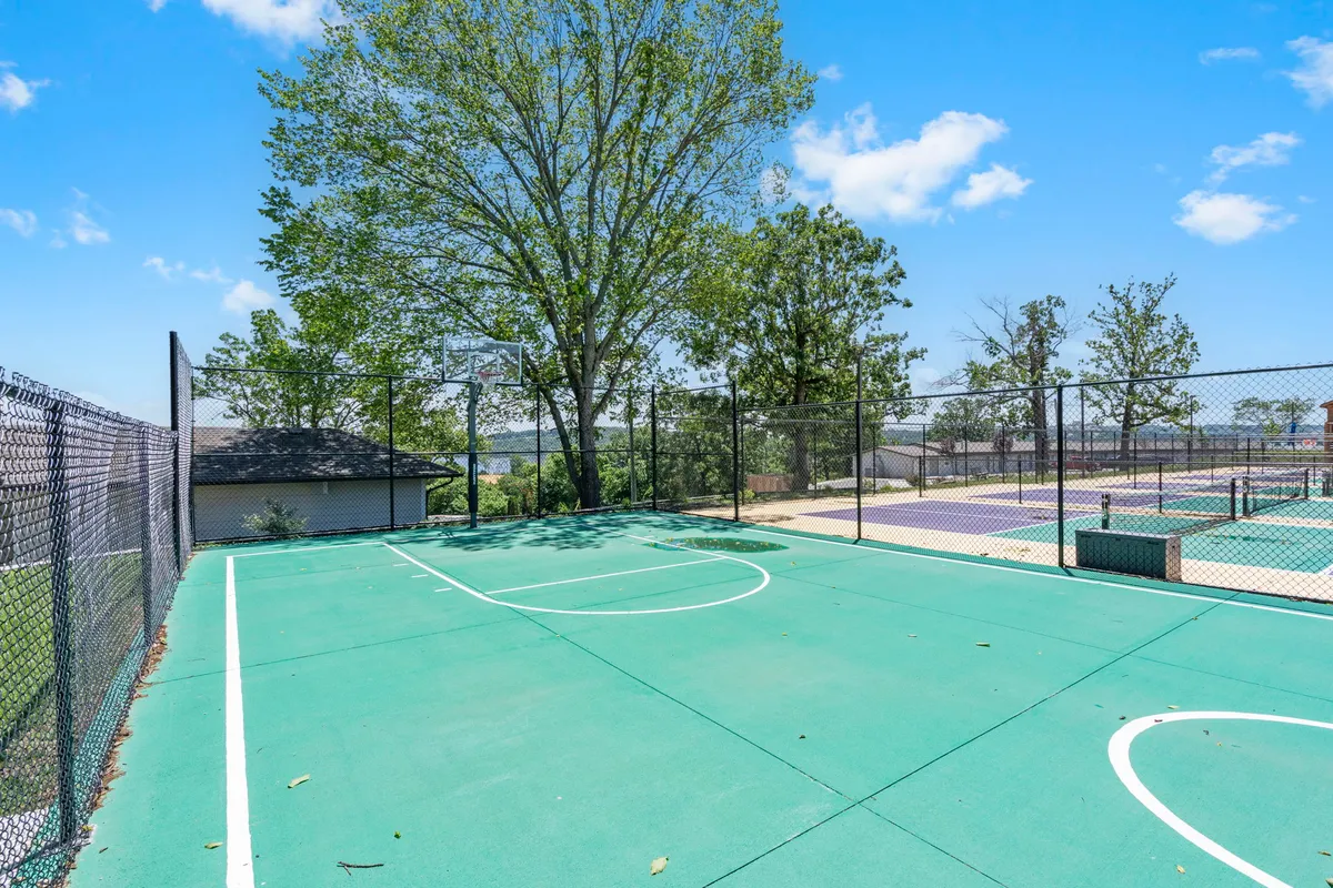 A lively exterior view of a basketball court surrounded by trees, with a clear green surface and a prominent basketball hoop. The open space invites energetic play, group games, and outdoor exercise within the community setting.