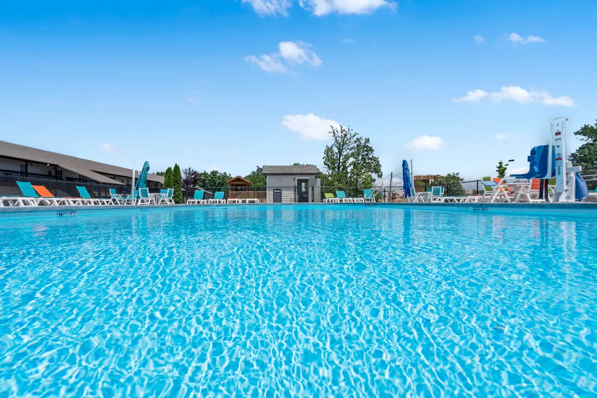 An outdoor pool area surrounded by lush greenery, with plenty of lounge chairs, a playground structure nearby, and a fun pool slide. Families and children are seen swimming, sunbathing, and playing, illustrating active enjoyment of the community's outdoor spaces.