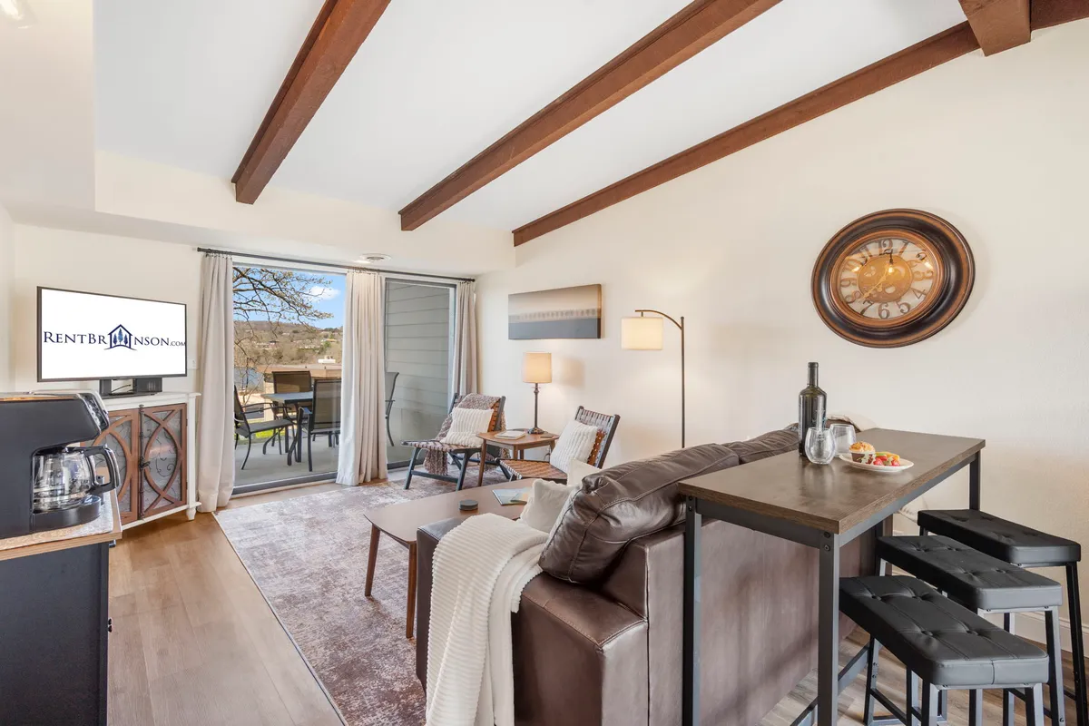 This image showcases a cozy living area with a dining table, a TV, a coffee maker, and a bright view outside through glass doors. The space is bathed in natural light, inviting relaxation and socializing. The furniture arrangement encourages group gatherings for meals, games, or TV watching.
