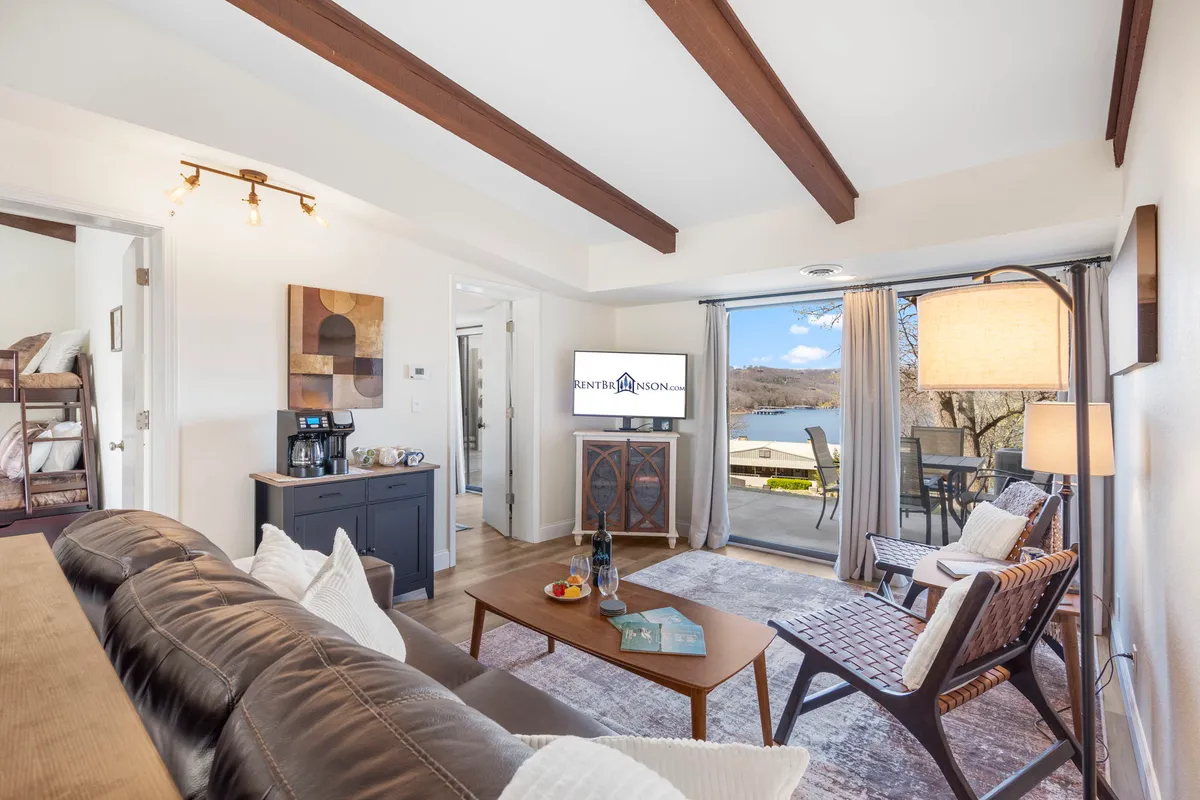 A casual, inviting living room setup with a large sofa, dining table, TV with branding, and a coffee station, all with a beautiful view of the lake outside. The setting suggests flexibility for dining, relaxing, socializing, or enjoying entertainment together.