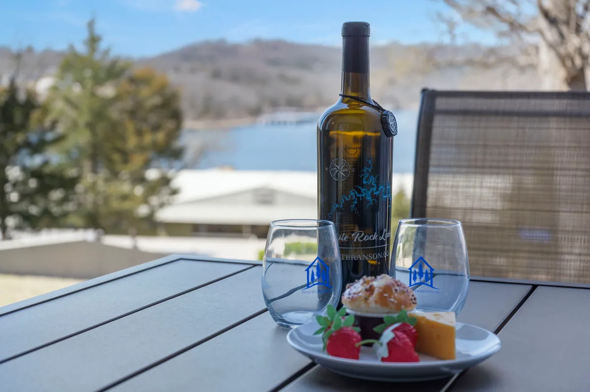 A table set with wine glasses, a bottle of wine, and a plate of dessert is positioned to overlook a tranquil lake. The background features distant hills and a shimmering water view, evoking leisurely, celebratory evenings outdoors.