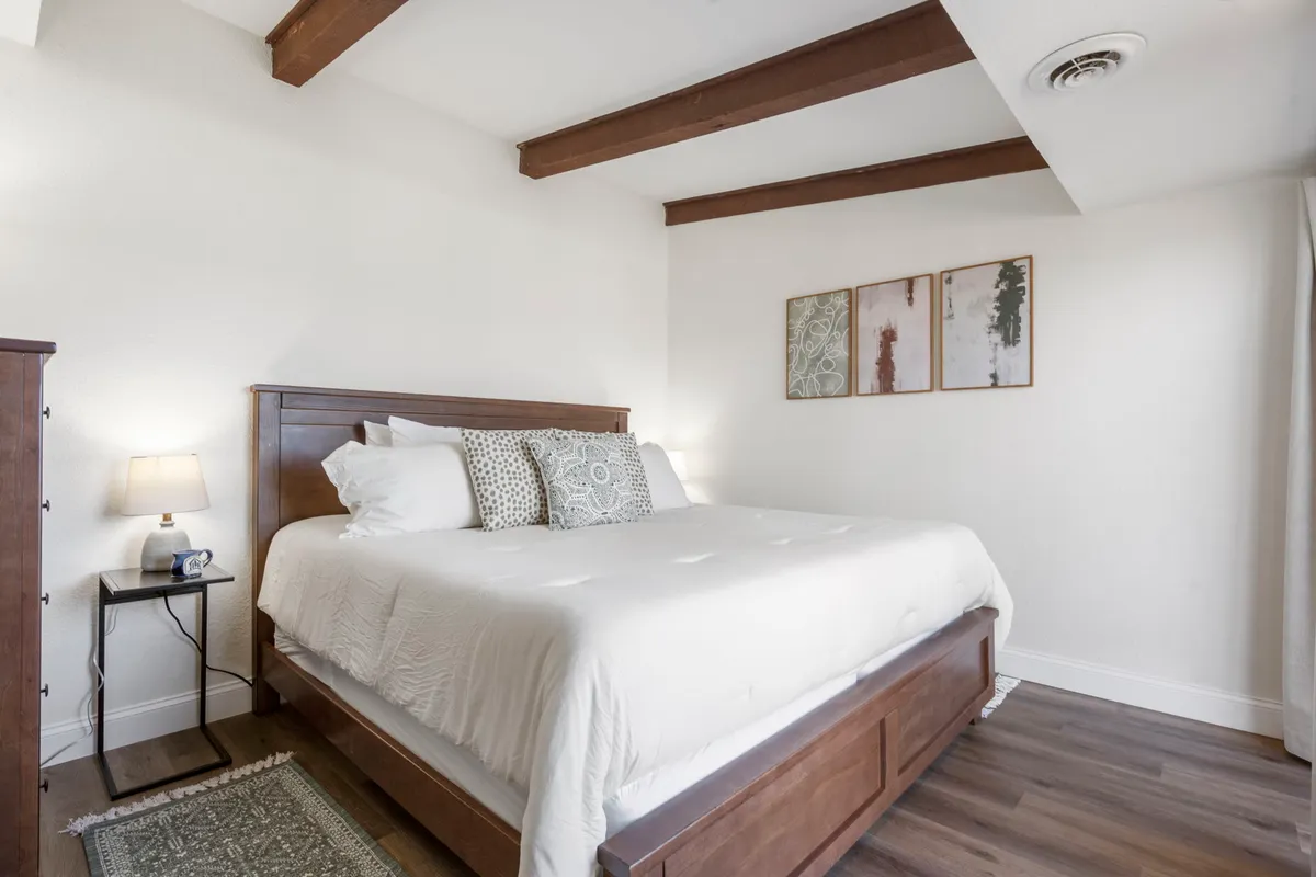 A tranquil bedroom featuring a plush bed with crisp white linens, decorative pillows, warm wooden beams on the ceiling, soft ambient lighting from a bedside lamp, and simple artwork on the wall. The room exudes a serene, restful atmosphere.