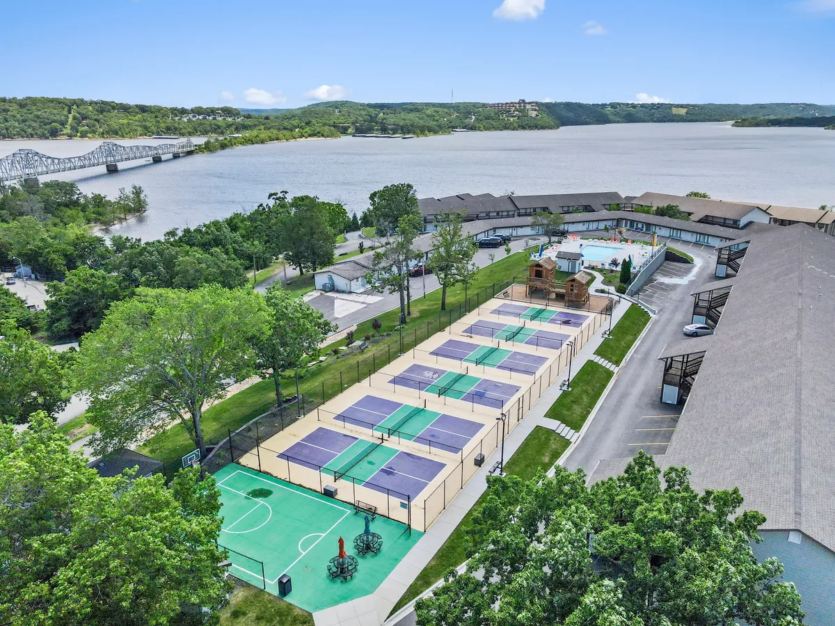 A wide-angle exterior shot capturing tennis courts, a basketball area, a swimming pool, surrounding greenery, and a partial waterfront view. Multiple active areas are visible, illustrating options for recreation and relaxation.
