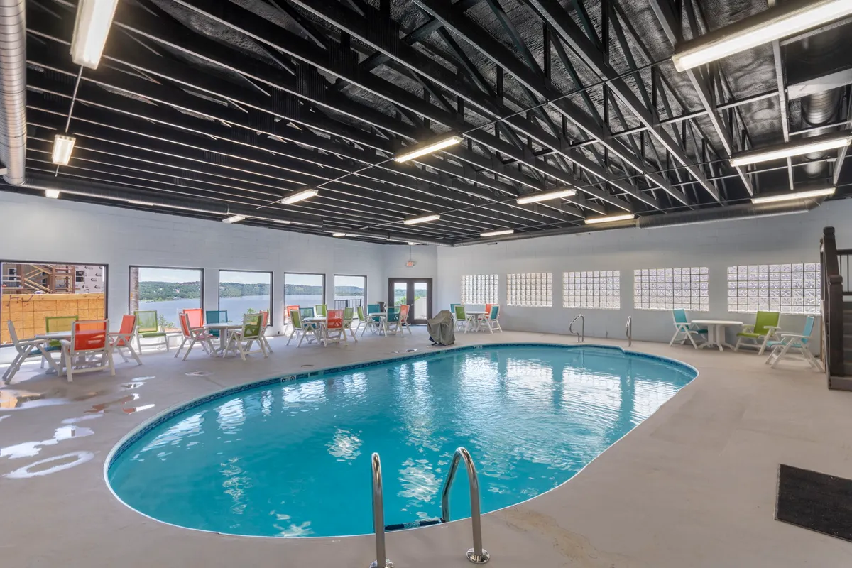 A bright, modern indoor swimming pool area featuring colorful lounge chairs and large windows that let in plenty of natural light, creating an inviting atmosphere for year-round swimming and relaxation. Groups of people are seen swimming, relaxing, and socializing by the poolside.